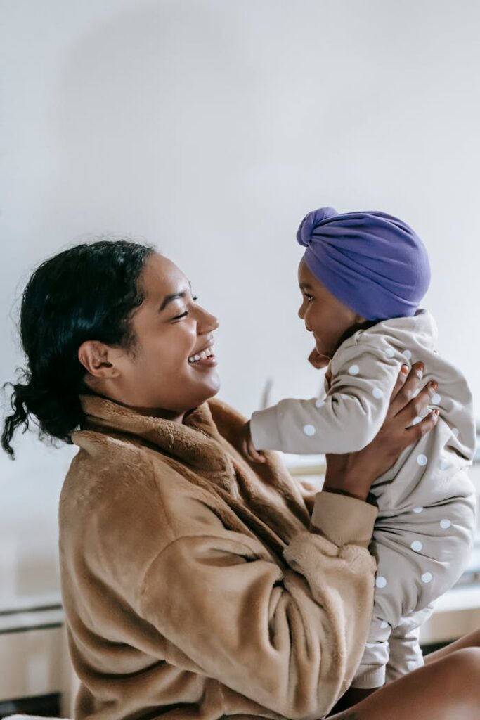 Side view of positive African American woman with little black baby in hands looking at each other in light room